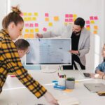 Pessoas analisando dados em uma tela grande de computador. Ao fundo, uma parede com post its coloridos