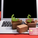 Imagem de um computador com caixas de papelão abertas sobre o teclado, com um cartão de crédito e cifrões