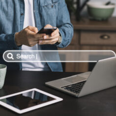 Homem de pé digitando no celular à frente de uma mesa com notebook e tablet, e barra de busca sobreposta à foto