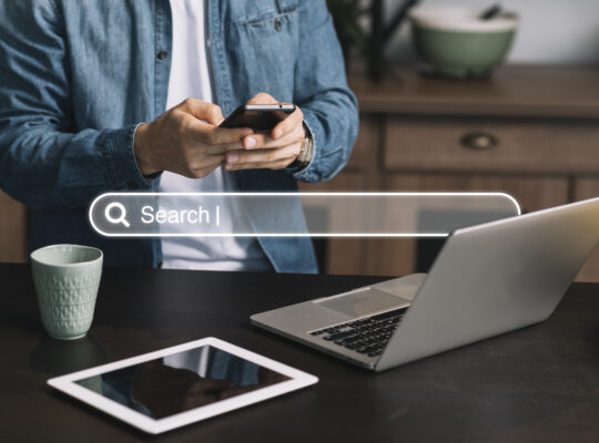 Homem de pé digitando no celular à frente de uma mesa com notebook e tablet, e barra de busca sobreposta à foto