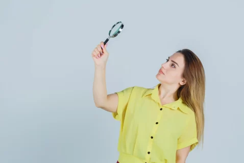 Mulher de camisa amarela usando uma lupa para encontrar produtos para vender em fundo branco
