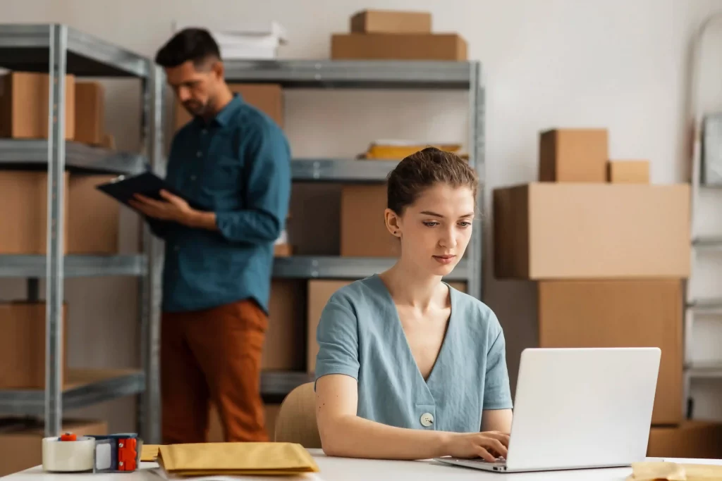 Mulher sentada em frente a um notebook com um homem ao fundo mexendo no estoque do Mercado Livre