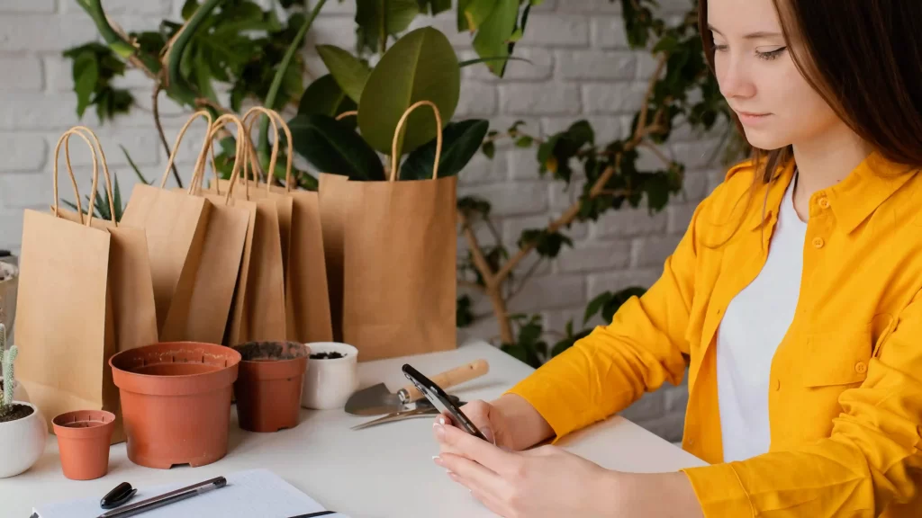 Mulher usando um celular sentada em uma cadeira a frente de uma mesa com sacolas sustentáveis e plantas ao fundo