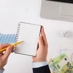 Homem segurando bloco e caneta com dinheiro e outros itens ao fundo