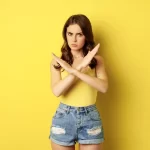 Mulher de camiseta amarela e short jeans fazendo sinal de X com os braços em fundo amarelo