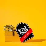 Embalagem de presente amarela com laço dourado ao lado de uma tag preta da Black Friday em fundo amarelo