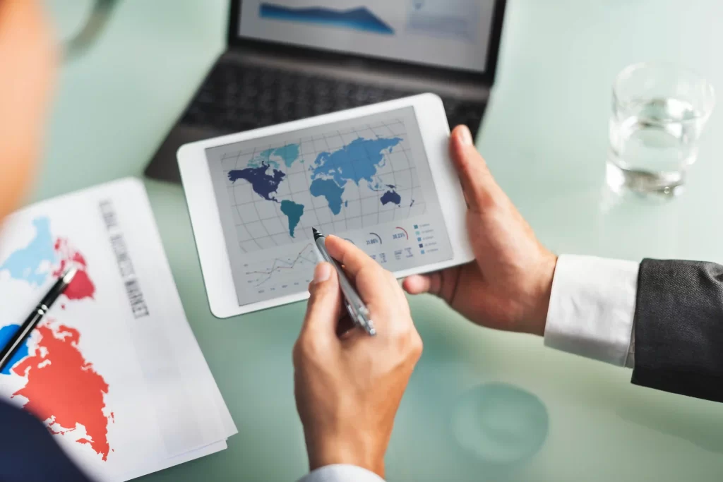Duas pessoas segurando um tablet com mapa do mundo ao lado de notebook e outros itens