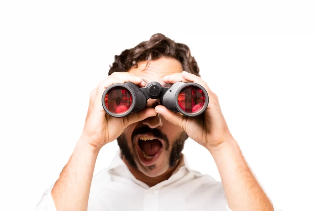 Homem segurando um binóculo com lentes vemelhas em frente aos seus olhos em fundo branco