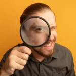 Homem com lupa em frente ao rosto em fundo amarelo