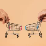 Duas mãos segurando carrinhos de compra de cores diferentes, um de frente para o outro em fundo laranja