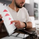 Homem de fone de ouvido segurando uma prancheta com gráficos apresentando para outra pessoa