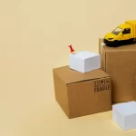 Caixas brancas e de papelão no chão com um caminhão amarelo em cima e alguns marcadores vermelhos em fundo amarelo