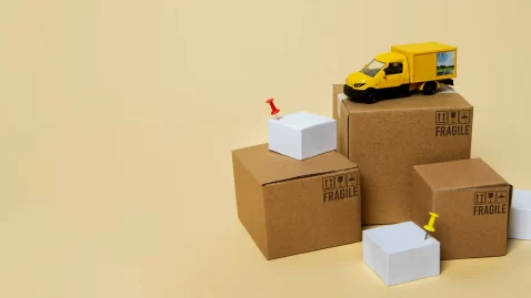 Caixas brancas e de papelão no chão com um caminhão amarelo em cima e alguns marcadores vermelhos em fundo amarelo