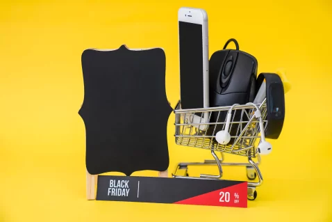 Carrinho de compras com três itens ao lado de um quadro negro e uma placa escrito Black Friday em fundo amarelo