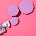 Mão segurando megafone com círculos lilás em frente em fundo rosa