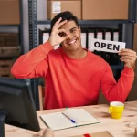 Homem sentado em mesa de escritório com uma mão no olho e outra segurando um papel escrito "open"