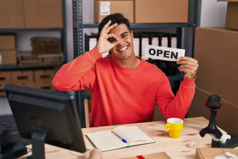 Homem sentado em mesa de escritório com uma mão no olho e outra segurando um papel escrito "open"