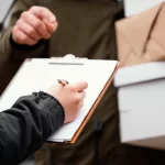 Mão de mulher segurando e escrevendo em uma prancheta logo a frente de um homem segurando caixas de entrega