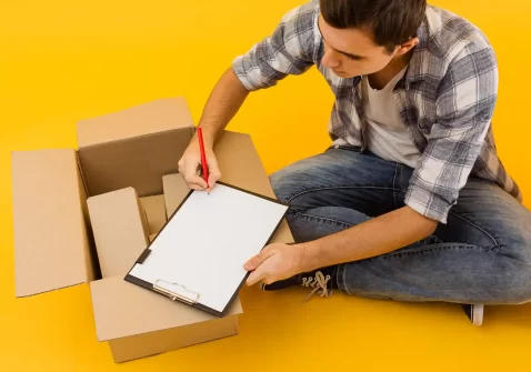 Jovem sentado no chão ao lado de caixas de papelão abertas, preenchendo uma prancheta, simbolizando os primeiros 90 dias vendendo no Mercado Livre