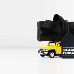 Miniatura de caminhão amarelo transportando presente embrulhado em preto com etiqueta Black Friday.
