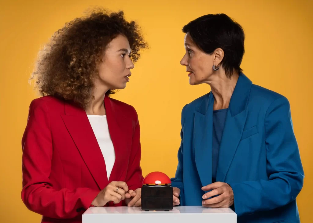 Duas mulheres de terno colorido discutem diante de botão vermelho sobre mesa branca, em fundo amarelo