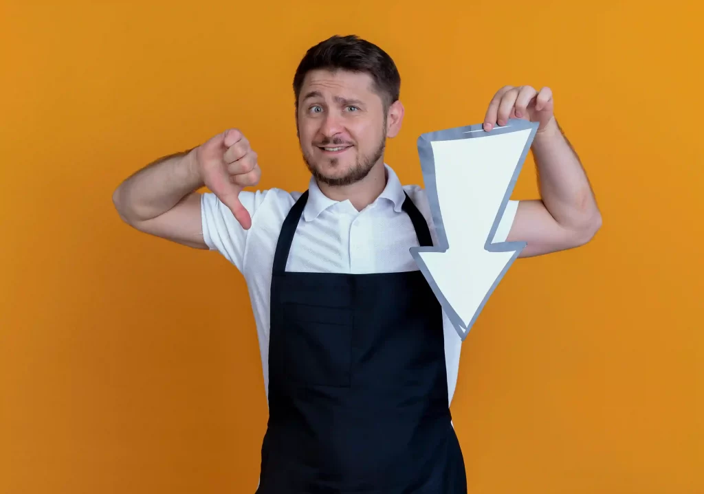 Homem de avental preto fazendo sinal negativo com uma mão e segurando uma seta para baixo com a outra em fundo amarelo