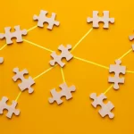 Várias peças de quebra-cabeças cinzas interligadas por um barbante amarelo em fundo laranja