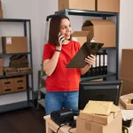Empreendedora em depósito organizando pedidos com prancheta, caixas e estoque de e-commerce.