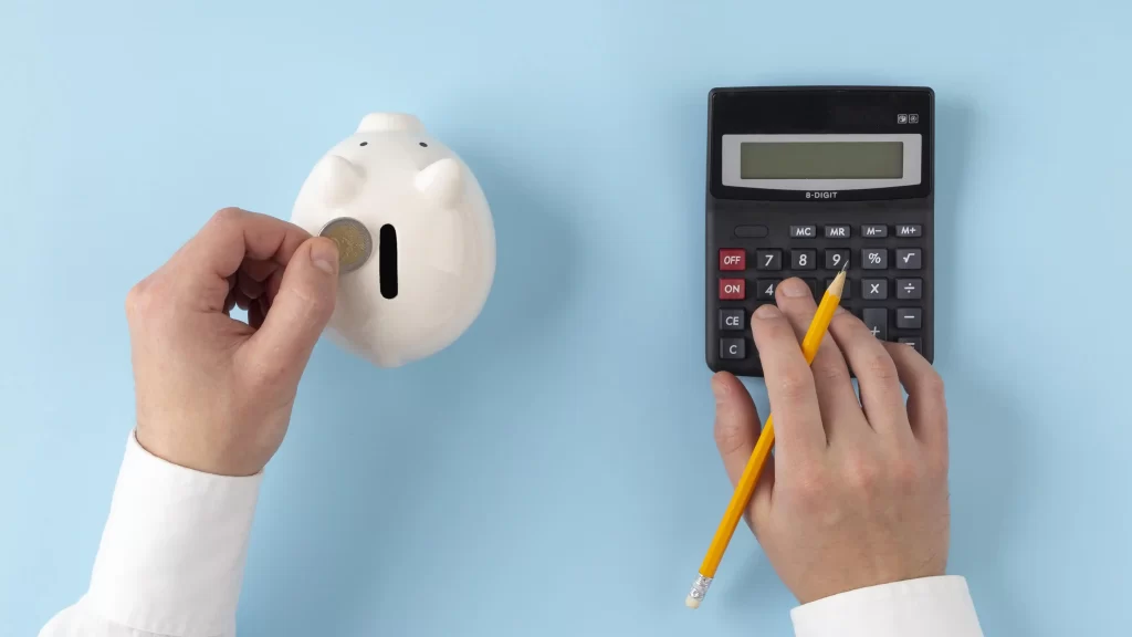 Mão inserindo moeda em cofre de porquinho branco ao lado de uma calculadora sobre fundo azul, simbolizando economia e gestão de micro-nichos rentáveis