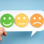 Mão segurando balão de fala com três emojis de avaliação: feliz, neutro e triste.