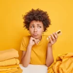 Mulher em balcão de loja com várias roupas amarelas sobre ele segurando um celular com uma mão e a outra no queixo.