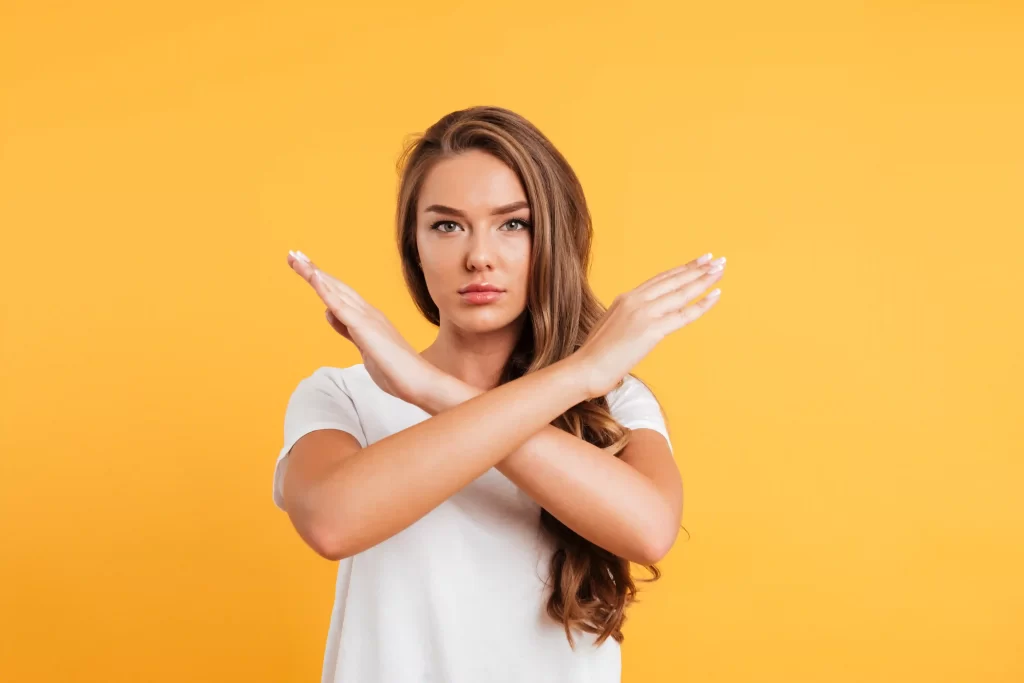 Mulher de camiseta branca em fundo amarelo fazendo gesto de X com os braços em sinal de recusa