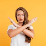 Mulher de camiseta branca em fundo amarelo fazendo gesto de X com os braços em sinal de recusa