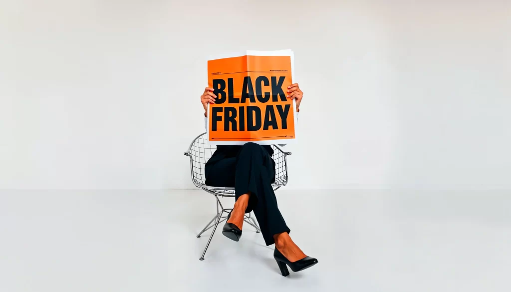 Pessoa sentada em uma cadeira com as pernas cruzadas, segurando um jornal laranja com o texto Black Friday em letras grandes