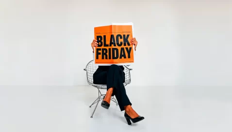 Pessoa sentada em uma cadeira com as pernas cruzadas, segurando um jornal laranja com o texto Black Friday em letras grandes