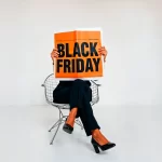 Pessoa sentada em cadeira com as pernas cruzadas segurando um jornal grande com o título Black Friday em destaque