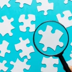 Lupa focando em uma peça de quebra-cabeça branca entre várias outras sobre fundo azul, representando o processo de analisar concorrentes e encontrar oportunidades