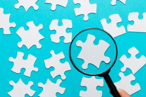 Lupa focando em uma peça de quebra-cabeça branca entre várias outras sobre fundo azul, representando o processo de analisar concorrentes e encontrar oportunidades