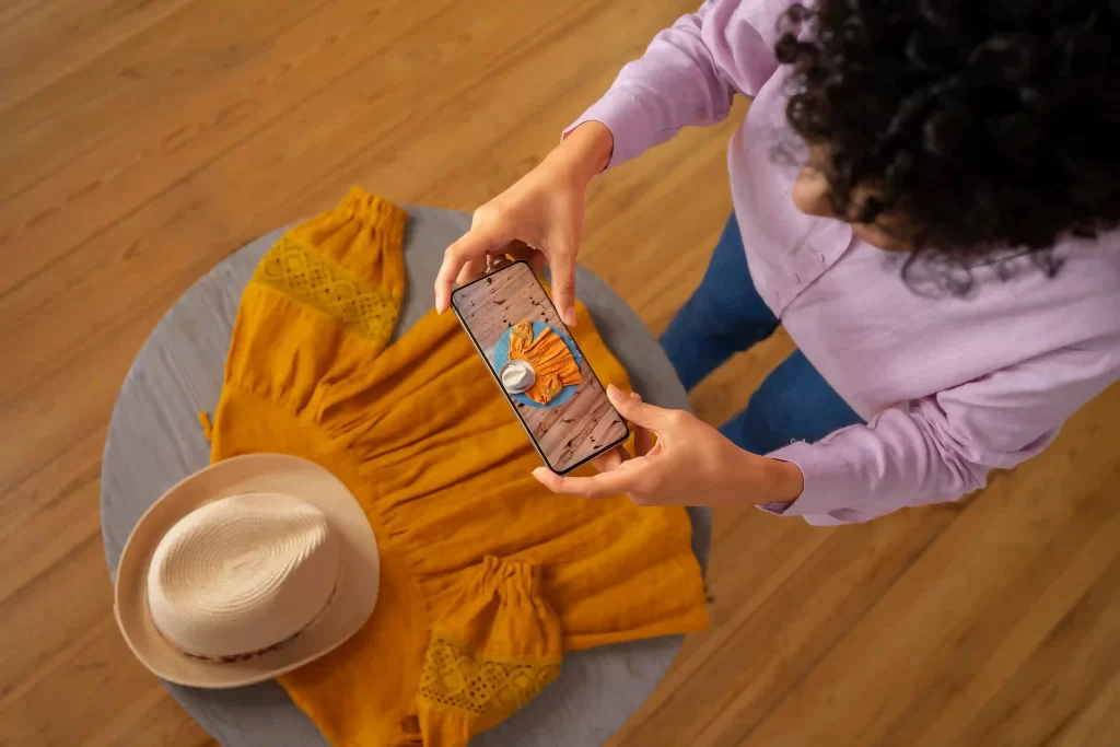 Pessoa fotografando com smartphone uma composição de roupa amarela e chapéu sobre a mesa, representando produção de conteúdo interativo para e-commerce de moda