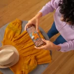 Pessoa fotografando com smartphone uma composição de roupa amarela e chapéu sobre a mesa, representando produção de conteúdo interativo para e-commerce de moda