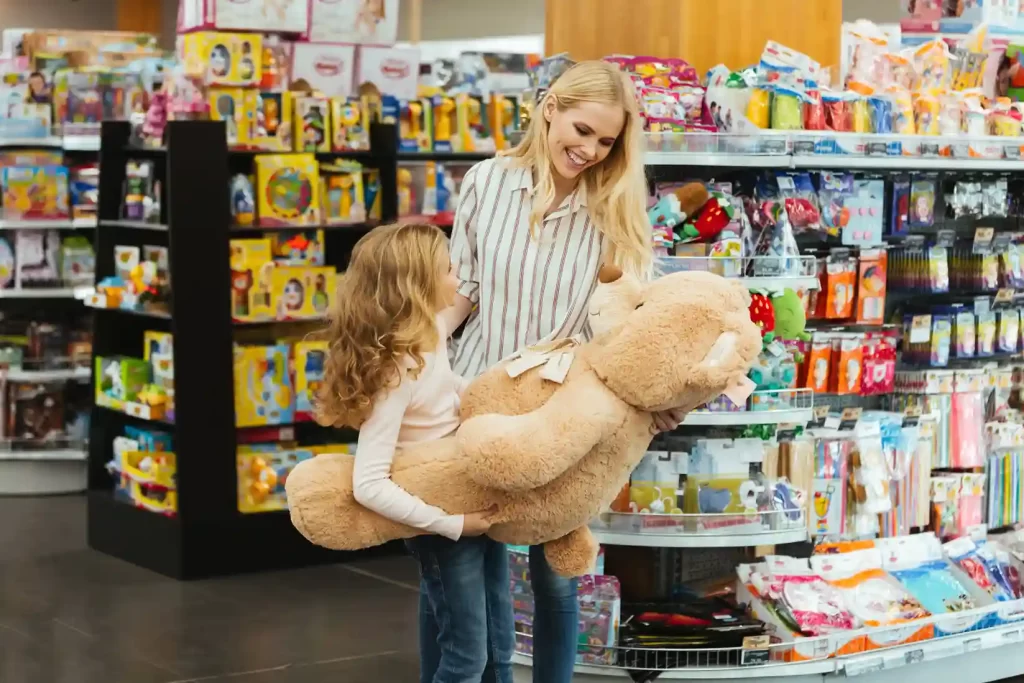 Mãe e filha em loja de brinquedos, sorrindo enquanto seguram um urso de pelúcia gigante, cercadas por prateleiras coloridas cheias de produtos infantis