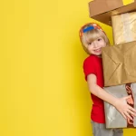 Criança sorridente de boné e camiseta vermelha segura várias caixas de presente embrulhadas em papel dourado e prateado