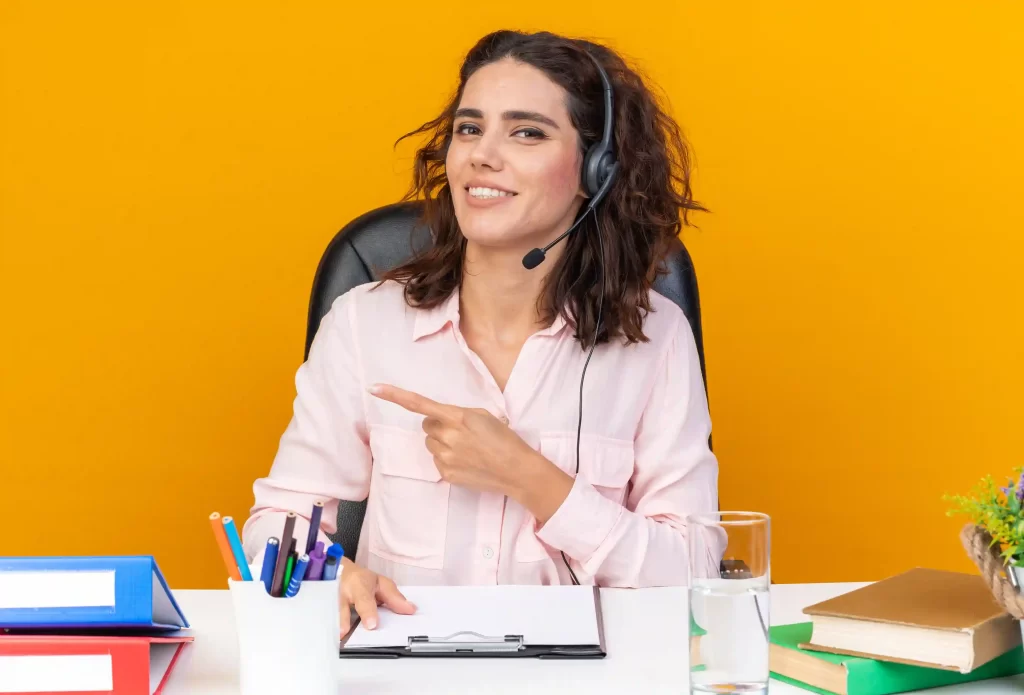 Mulher sorridente usando headset e apontando para o lado, sentada em mesa de escritório com papéis e canetas, representando atendimento ao cliente como parte da estratégia de pós-venda