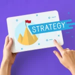 Mãos segurando tablet com ilustração de montanha e bandeira vermelha com a palavra strategy, simbolizando metas e planejamento dentro de uma estratégia de pós-venda