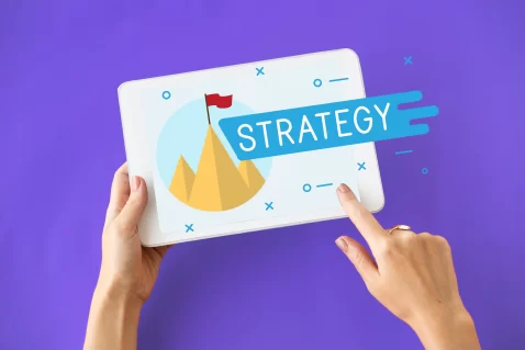 Mãos segurando tablet com ilustração de montanha e bandeira vermelha com a palavra strategy, simbolizando metas e planejamento dentro de uma estratégia de pós-venda