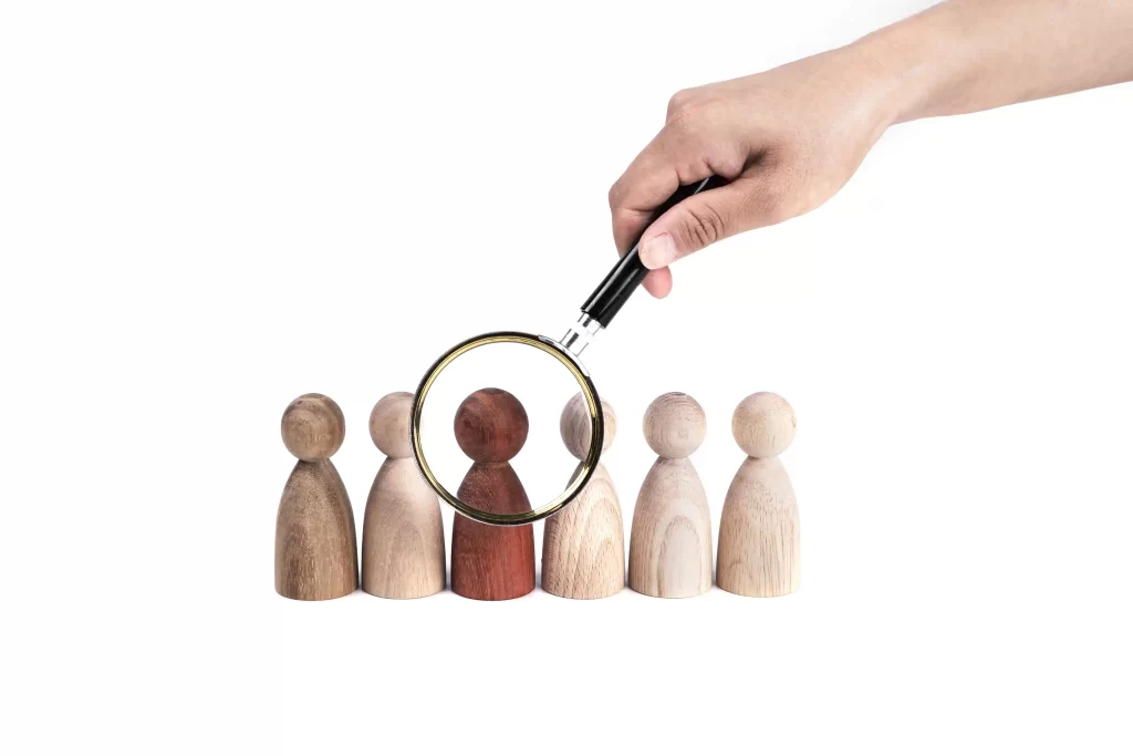 Mão segurando uma lupa sobre peças de madeira em formato de pessoas, destacando uma delas, simbolizando o processo de identificar concorrentes indiretos no mercado