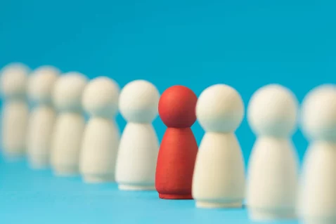 Peças de madeira alinhadas com uma figura vermelha destacada entre figuras brancas, representando diferenciação e identificação de concorrentes indiretos