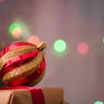Bola de Natal vermelha e dourada apoiada sobre um presente embrulhado, com luzes coloridas desfocadas ao fundo, simbolizando o espírito do Natal 2025 e o clima de consumo