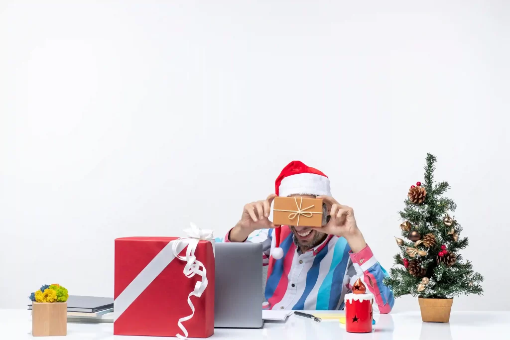 Homem usando gorro de Papai Noel segurando uma caixa de presente em frente ao notebook, com árvore de Natal e pacotes ao redor
