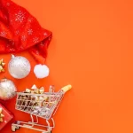 Carrinho de compras em miniatura com laços dourados e bolas de Natal sobre fundo laranja, representando a antecipação de tendências e oportunidades de venda no Natal